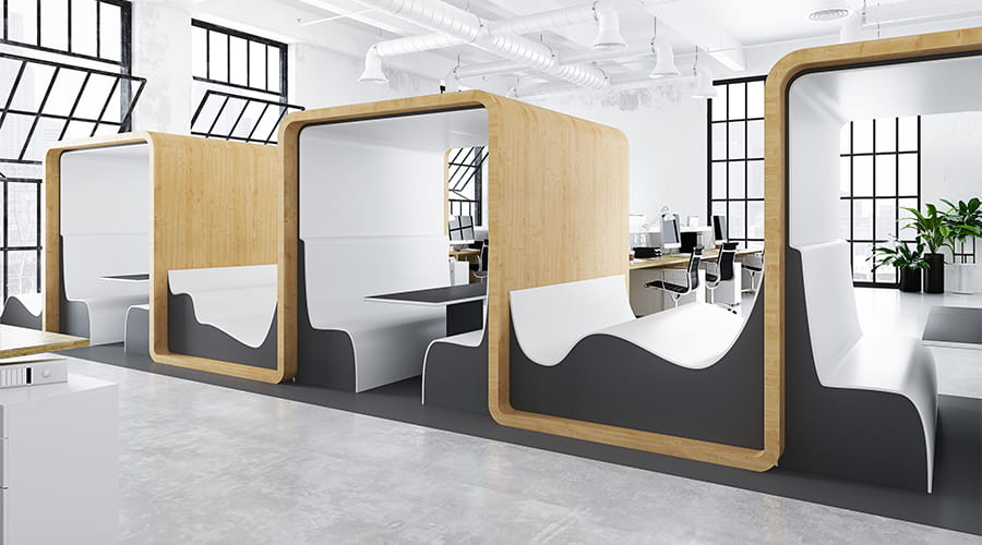 Interno di ufficio open space moderno con cabine in legno dal design arrotondato, dotate di scrivania e sedute integrate, in ambiente luminoso con grandi finestre industriali e arredi contemporanei.