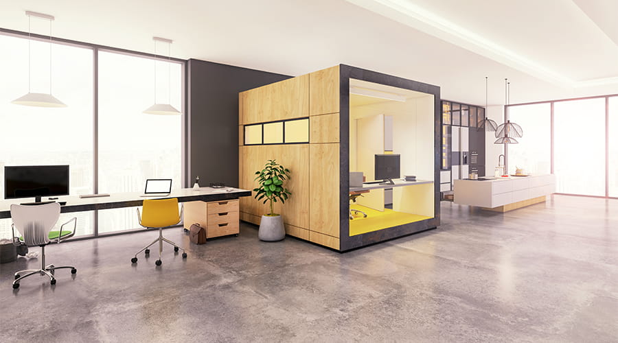 Spazio ufficio moderno e luminoso con postazioni di lavoro, pavimento in cemento, grande box in legno e vetro per riunioni o ufficio privato, e area cucina sullo sfondo con design minimalista.