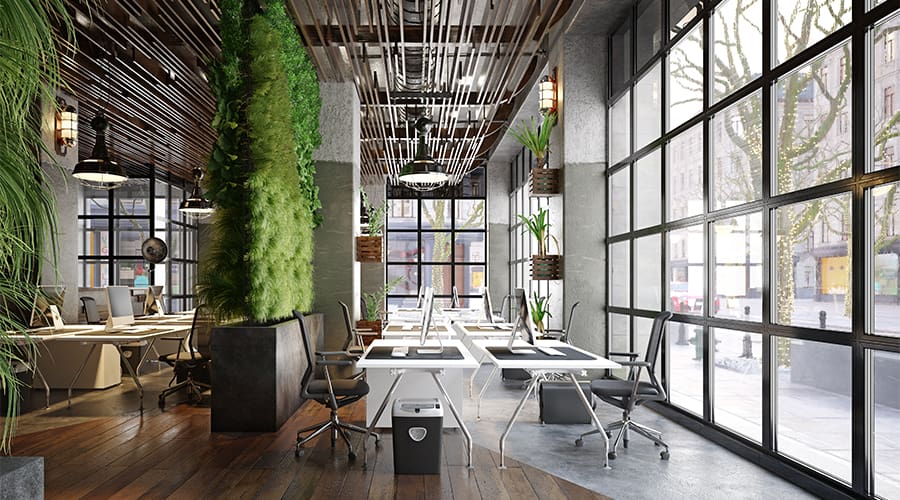 Spazio ufficio open space in stile industriale con scrivanie condivise, computer desktop, sedie ergonomiche, parete verde verticale con piante e ampie finestre a tutta altezza che affacciano sulla città.