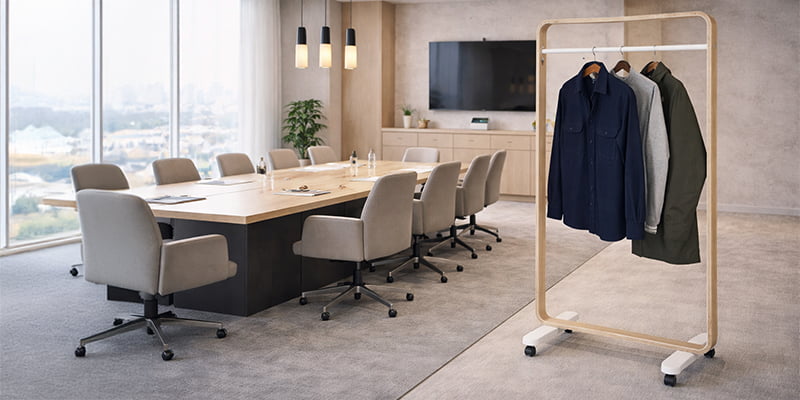 Stender appendiabiti con struttura in legno e barra in metallo con abiti appesi, posizionato in una moderna sala riunioni con tavolo conferenze e sedie di design.